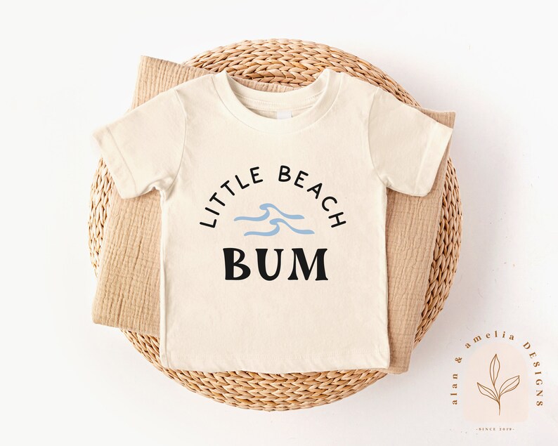 以下が含まれることがあります： 黒と青の文字で「Little Beach Bum」と書かれた白い半袖Tシャツ。テキストは青い波のグラフィックで囲まれています。
