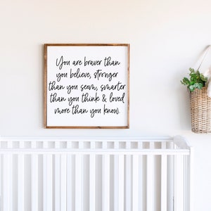 May include: A framed wooden sign with the text "You are braver than you believe, stronger than you seem, smarter than you think & loved more than you know." A white cot is in the foreground. A woven basket with a stuffed lamb hangs on the wall.