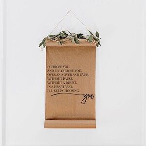 May include: A brown paper scroll with a black inscription that reads "I choose you. And I'll choose you, over and over and over. Without pause. Without a doubt. In a heartbeat. I'll keep choosing you." The scroll is hanging from a string with a garland of green leaves and white flowers.