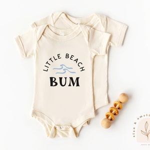以下が含まれることがあります： クリーム色の半袖ベビーロンパース。黒いグラフィックには「Little Beach Bum」の文字と青い波のデザインが描かれています。