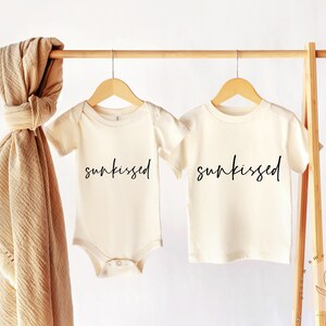 May include: Two cream-colored shirts with short sleeves hanging on a wooden rack. The shirts have the word "sunkissed" written in black cursive on the front.