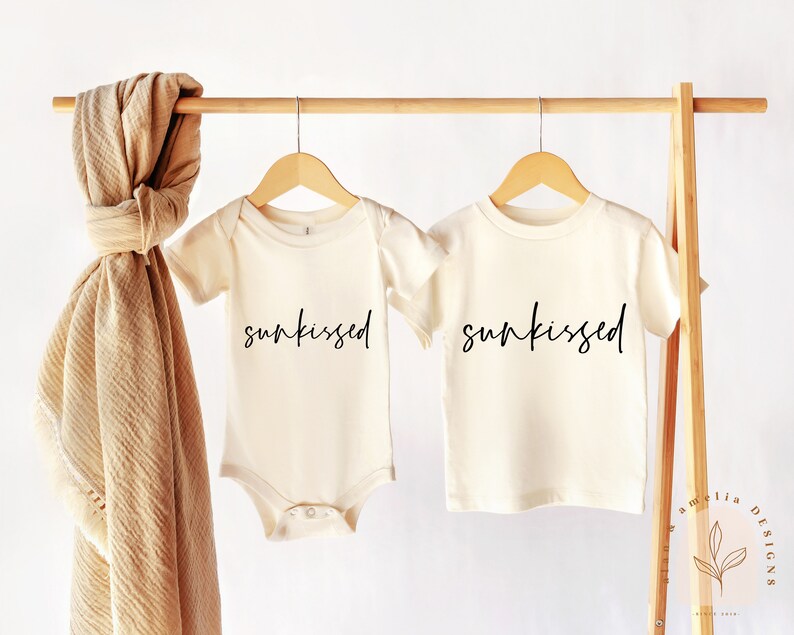 May include: Two cream-colored shirts with short sleeves hanging on a wooden rack. The shirts have the word "sunkissed" written in black cursive on the front.