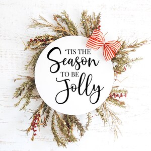 May include: White round sign with the text "'Tis the season to be jolly" in black cursive font. The sign is surrounded by a wreath of green branches with red berries and a red and white striped bow.
