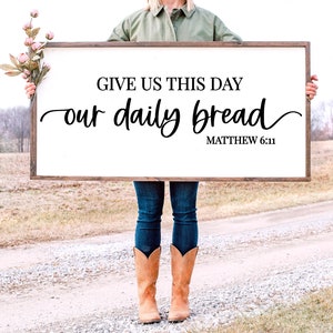 May include: A white wooden sign with the text "Give us this day our daily bread Matthew 6:11" in black lettering. The sign is being held by a person wearing blue jeans and brown cowboy boots.