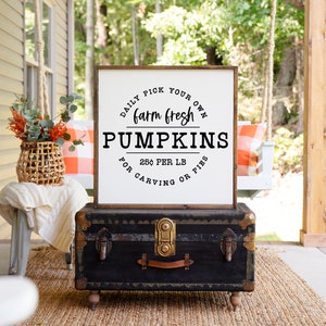May include: A white wooden sign with black text that reads "Daily Pick Your Own Farm Fresh Pumpkins 25¢ Per LB For Carving Or Pies". The sign is hanging on a porch with a wicker basket filled with autumn foliage and a black vintage trunk.