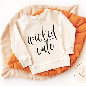 May include: A cream-colored sweatshirt with the text "wicked cute" in black lettering. The sweatshirt is on a light orange background.