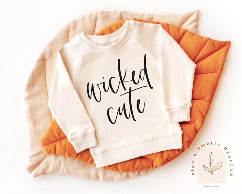 May include: A cream-colored sweatshirt with the text "wicked cute" in black lettering. The sweatshirt is on a light orange background.