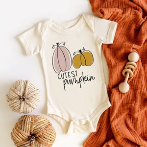 May include: A white baby onesie with a pink and yellow pumpkin design and the text "Cutest Pumpkin". The onesie is lying on a white surface with two woven pumpkin decorations and a brown blanket.