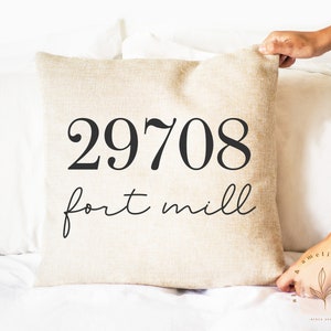 May include: A beige square cushion with the black text "29708 fort mill". The cushion is on a white surface. The cushion is being held by a person.