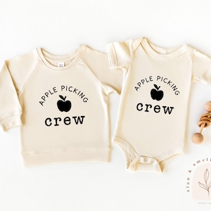 Peut inclure: Deux tenues de bébé couleur crème avec du texte noir qui dit "Apple Picking Crew" et un graphique de pomme noir.