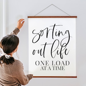 Puede incluir: Un cartel de pared de lona blanca con el texto "sorting out life one load at a time" en escritura cursiva negra. El cartel está siendo colgado en una pared por una persona.