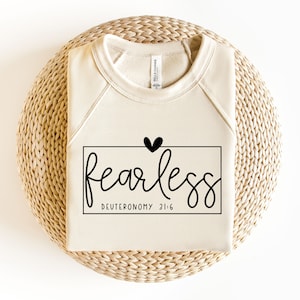 Puede incluir: Una sudadera beige con una caja negra que dice "fearless" con un pequeño corazón negro sobre la palabra y "DEUTERONOMY 31:6" debajo de la palabra.