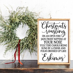 May include: A white wooden sign with black text that reads "Chestnuts roasting on an open fire Jack Frost nipping at your nose Yule tide carols being sung by a choir and folks dressed up like Eskimos". The sign is leaning against a white wall with a green wreath and red ribbon.