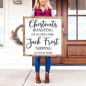 May include: A wooden sign with the text "Chestnuts Roasting on an Open Fire Jack Frost Nipping at Your Nose" in black calligraphy font. The sign is being held by a person in front of a wooden door.