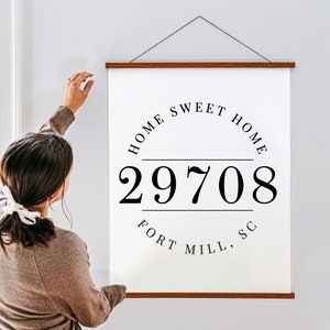 Może przedstawiać: Biały obraz na płótnie z napisem "HOME SWEET HOME" w okrągłym wzorze, wraz z numerem "29708" i "FORT MILL, SC" w kolorze czarnym. Płótno jest trzymane przez drewniane pręty.