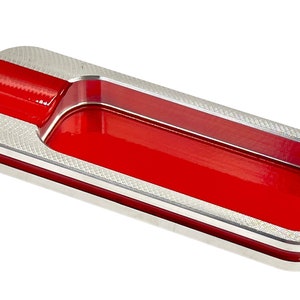 Op de afbeelding: Een rechthoekige asbak van zilver en rood met een verhoogde rand en een groef voor sigaretten. De asbak heeft een getextureerd oppervlak en een transparante kunststof inzet.