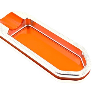 Könnte beinhalten: Aschenbecher in Form eines Bleistifts, orange und silberfarben. Der Aschenbecher hat eine tiefe Vertiefung zum Auffangen von Zigarettenstummeln.