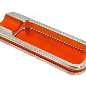 May include: A silver and orange rectangular ashtray with a groove for holding cigarettes. The ashtray has a smooth, polished surface and a modern design.