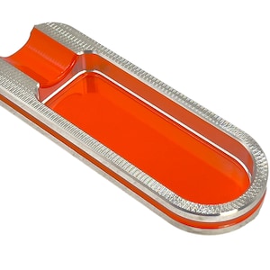 Könnte beinhalten: Ein rechteckiger Aschenbecher in Silber und Orange mit einem erhöhten Rand. Der Aschenbecher hat eine glatte, glänzende Oberfläche und einen strukturierten Rand.