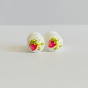 May include: Two white, round earrings with a painted strawberry design. Each earring features a bright red strawberry with green leaves and yellow flowers. The earrings are set against a plain white background.
