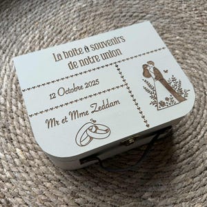 Peut inclure: Un coffret souvenir en bois clair avec texte et illustrations gravés. Le coffret porte l'inscription "La boîte à souvenirs de notre union", la date "12 Octobre 2025" et les noms "Mr et Mme Zeddam". Il y a aussi une illustration d'un couple et d'alliances.