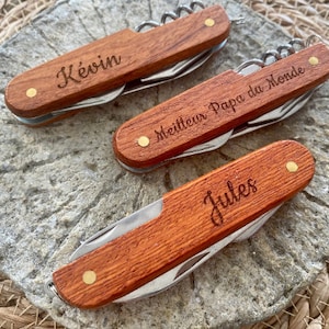 Peut inclure: Trois couteaux de poche en bois avec des noms gravés. Le premier couteau dit "Kevin", le deuxième dit "Meilleur Papa du Monde", et le troisième dit "Jules".