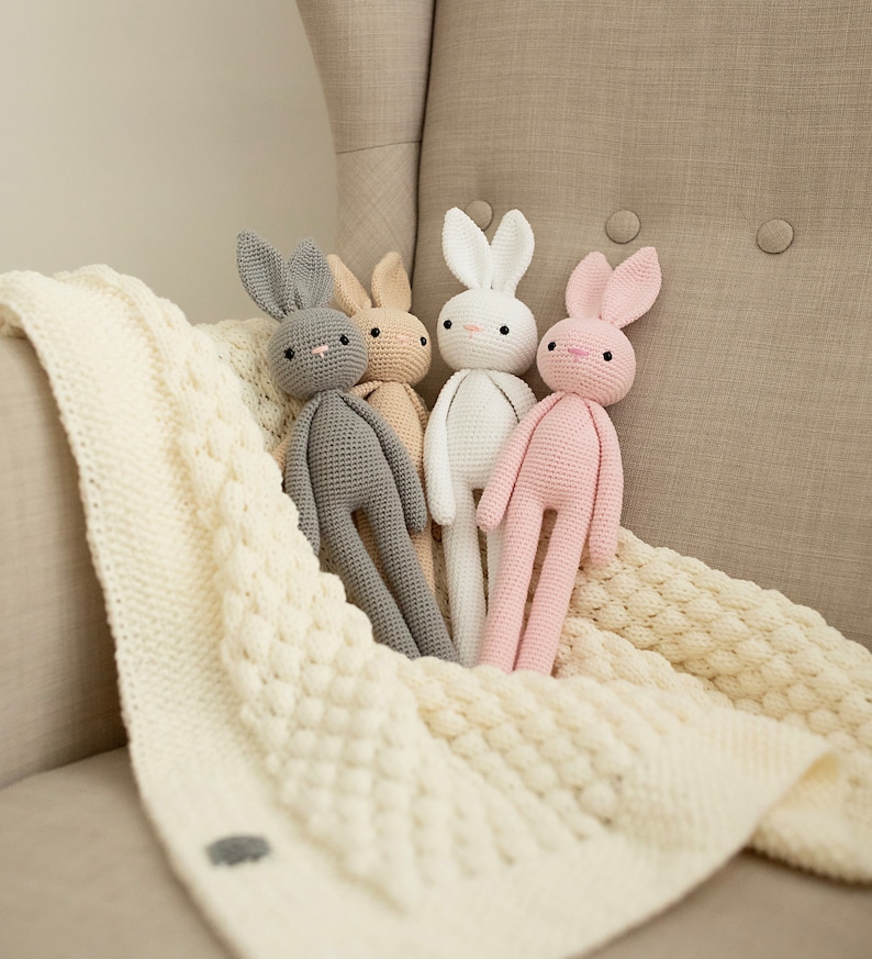 May include: Four crocheted bunny toys in different colors: gray, beige, white, and pink. The bunnies are sitting on a white knitted blanket.
