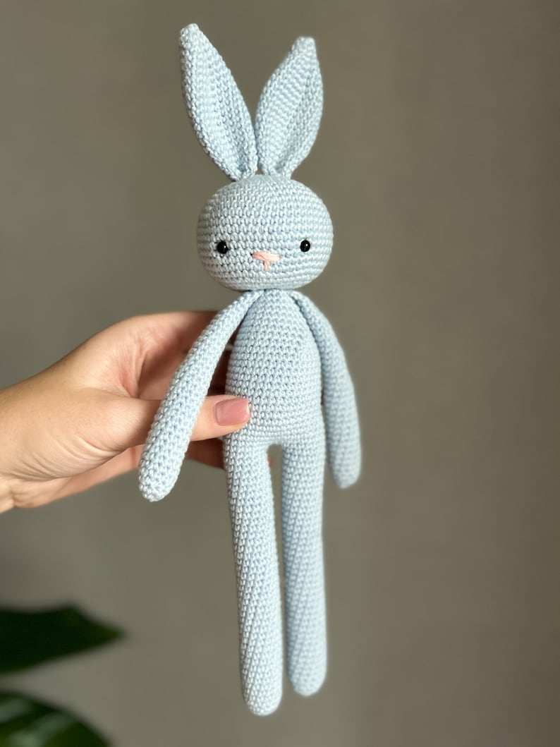May include: A light blue crocheted bunny rabbit toy with black eyes and a pink nose. The bunny has long arms and legs.