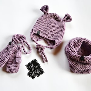 May include: A set of knitted baby accessories in a light purple color. The set includes a hat with ear flaps, a pair of mittens, and a neck warmer. The hat has a tie at the back. The accessories are made with a chunky yarn and have a simple, classic design.
