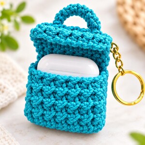 May include: A bright turquoise crocheted case designed to hold wireless earbuds, featuring a textured stitch pattern, a small top handle, and a gold-toned metal keychain ring attached to the side. The case is open, revealing a white earbud charging case.