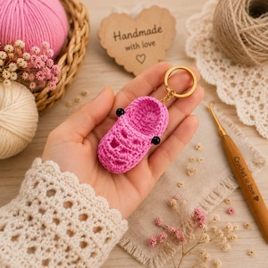 May include: A bright pink crocheted clog keychain featuring a gold-toned metal ring. The miniature shoe is crafted with detailed stitching, including small ventilation holes and two black circular accents on the sides, mimicking a classic clog design.