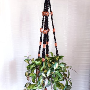 May include: A black and brown macrame plant hanger, with a wooden ring at the top. The plant hanger is holding a potted plant with green and variegated leaves. The design incorporates decorative wooden beads. The plant hanger is handmade.