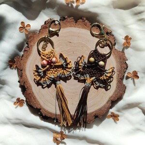 May include: Two owl-shaped keychains made with macrame and beads. The owl on the left has yellow, brown, and gray yarn with red and white beads for eyes. The owl on the right has brown, gray, and white yarn with brown and green beads for eyes. Both owls have tassels.