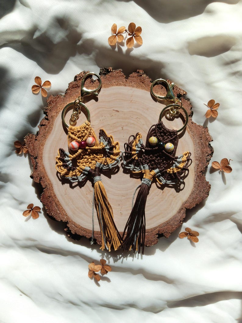 May include: Two owl-shaped keychains made with macrame and beads. The owl on the left has yellow, brown, and gray yarn with red and white beads for eyes. The owl on the right has brown, gray, and white yarn with brown and green beads for eyes. Both owls have tassels.