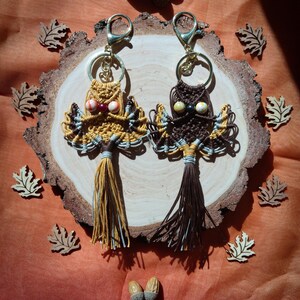 May include: Two owl-shaped macrame keychains with brown and yellow tassels hanging on a wooden slice. The owls have brown, white, and yellow string details and colorful bead eyes.