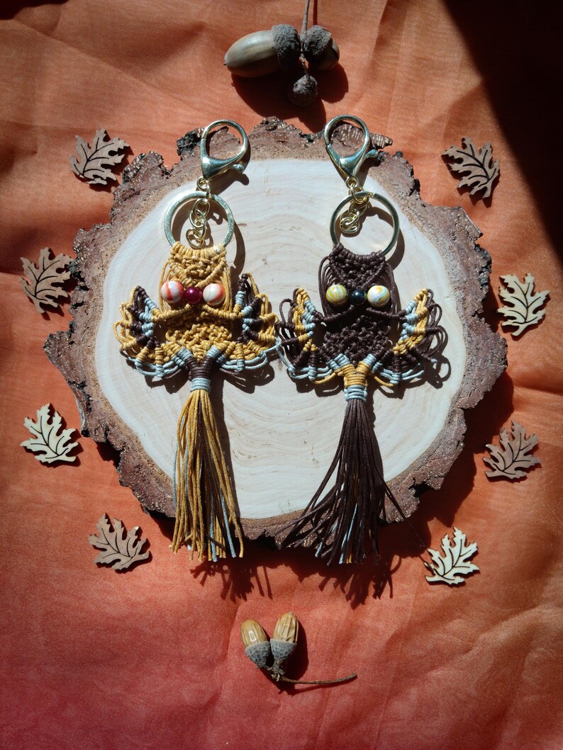 May include: Two owl-shaped macrame keychains with brown and yellow tassels hanging on a wooden slice. The owls have brown, white, and yellow string details and colorful bead eyes.