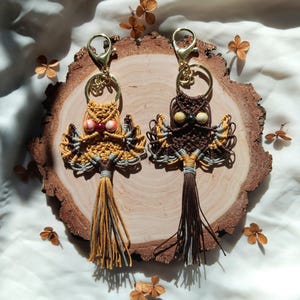 May include: Two owl-shaped keychains made with macrame and beads. The owls are brown and yellow with gray accents. The keychains are hanging from a wooden slice.