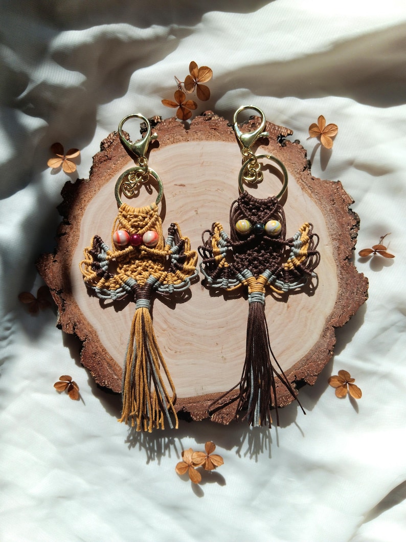 May include: Two owl-shaped keychains made with macrame and beads. The owls are brown and yellow with gray accents. The keychains are hanging from a wooden slice.