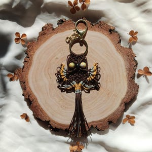 May include: A brown and yellow macrame owl keychain with a gold key ring. The owl has bead eyes and a brown tassel.