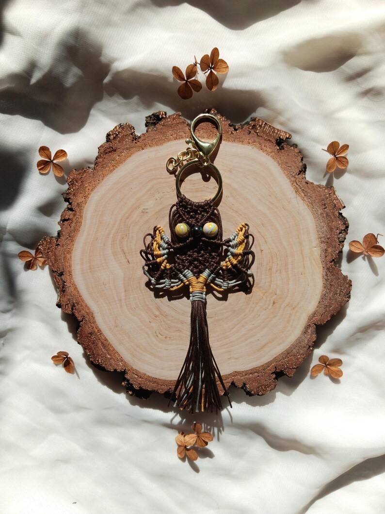 May include: A brown and yellow macrame owl keychain with a gold key ring. The owl has bead eyes and a brown tassel.