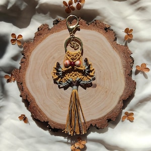 May include: A yellow and gray macrame owl keychain with a red and white bead for an eye. The keychain is hanging from a gold metal ring and is on a wooden slice.