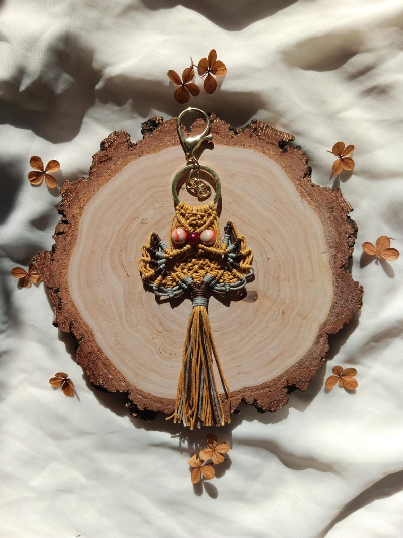 May include: A yellow and gray macrame owl keychain with a red and white bead for an eye. The keychain is hanging from a gold metal ring and is on a wooden slice.