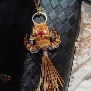 May include: A brown and white macrame owl keychain with a red and white bead for eyes. The keychain is attached to a gold key ring and is hanging from a black purse.