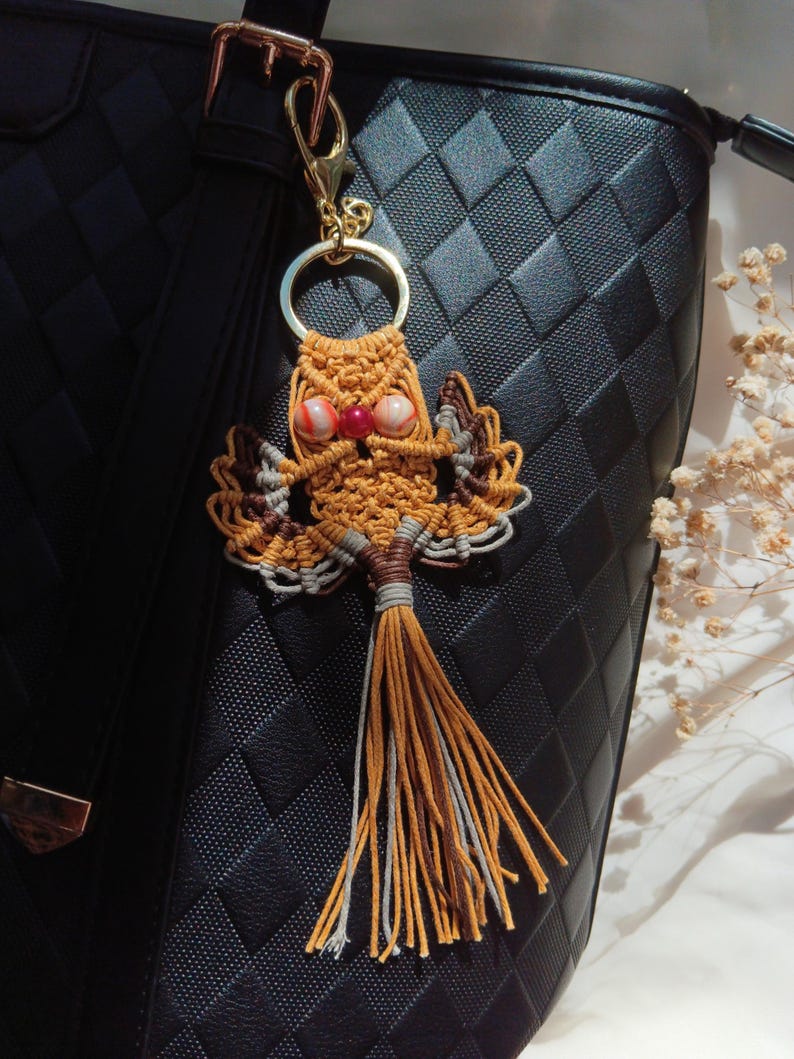 May include: A brown and white macrame owl keychain with a red and white bead for eyes. The keychain is attached to a gold key ring and is hanging from a black purse.