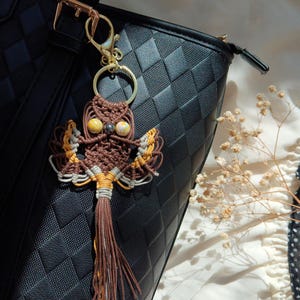 May include: A brown and yellow owl keychain with a tassel hanging from a black leather bag. The owl is made of macrame and has bead eyes.