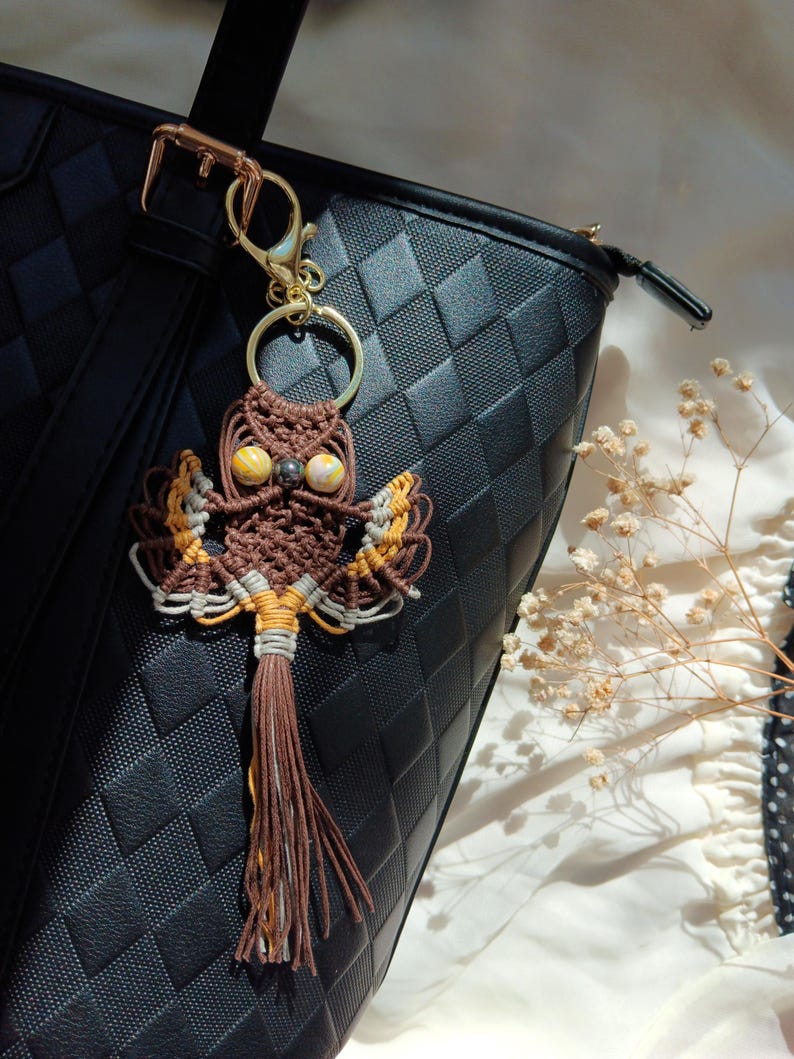 May include: A brown and yellow owl keychain with a tassel hanging from a black leather bag. The owl is made of macrame and has bead eyes.