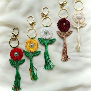 Könnte beinhalten: Eine Sammlung handgefertigter Blumen-Schlüsselanhänger in verschiedenen Farben, darunter Rot, Gelb, Weiß und Burgunder. Jeder Schlüsselanhänger hat einen goldfarbenen Verschluss und Ring, mit Quaste und grünen Blättern. Auf weißem Stoffhintergrund.