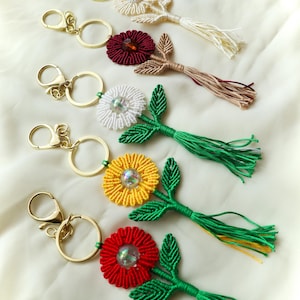Peut inclure: Plusieurs porte-clés fleuris faits à la main, de couleurs variées : blanc, bordeaux, jaune et rouge. Chaque porte-clés est doté d'un fermoir et d'un anneau dorés, de feuilles vertes et d'un pompon. Les fleurs ont un centre perlé.