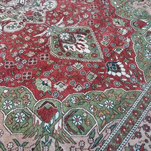 May include: Close-up of a vintage rug with a detailed floral pattern. The rug features a central field of deep red, bordered by green and beige sections. Intricate floral and geometric designs are woven throughout the rug's surface.
