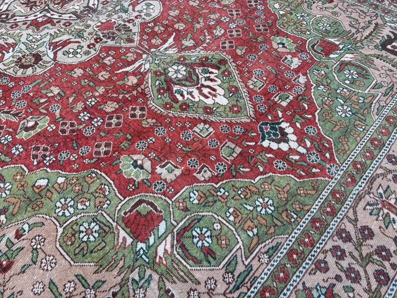 May include: Close-up of a vintage rug with a detailed floral pattern. The rug features a central field of deep red, bordered by green and beige sections. Intricate floral and geometric designs are woven throughout the rug's surface.